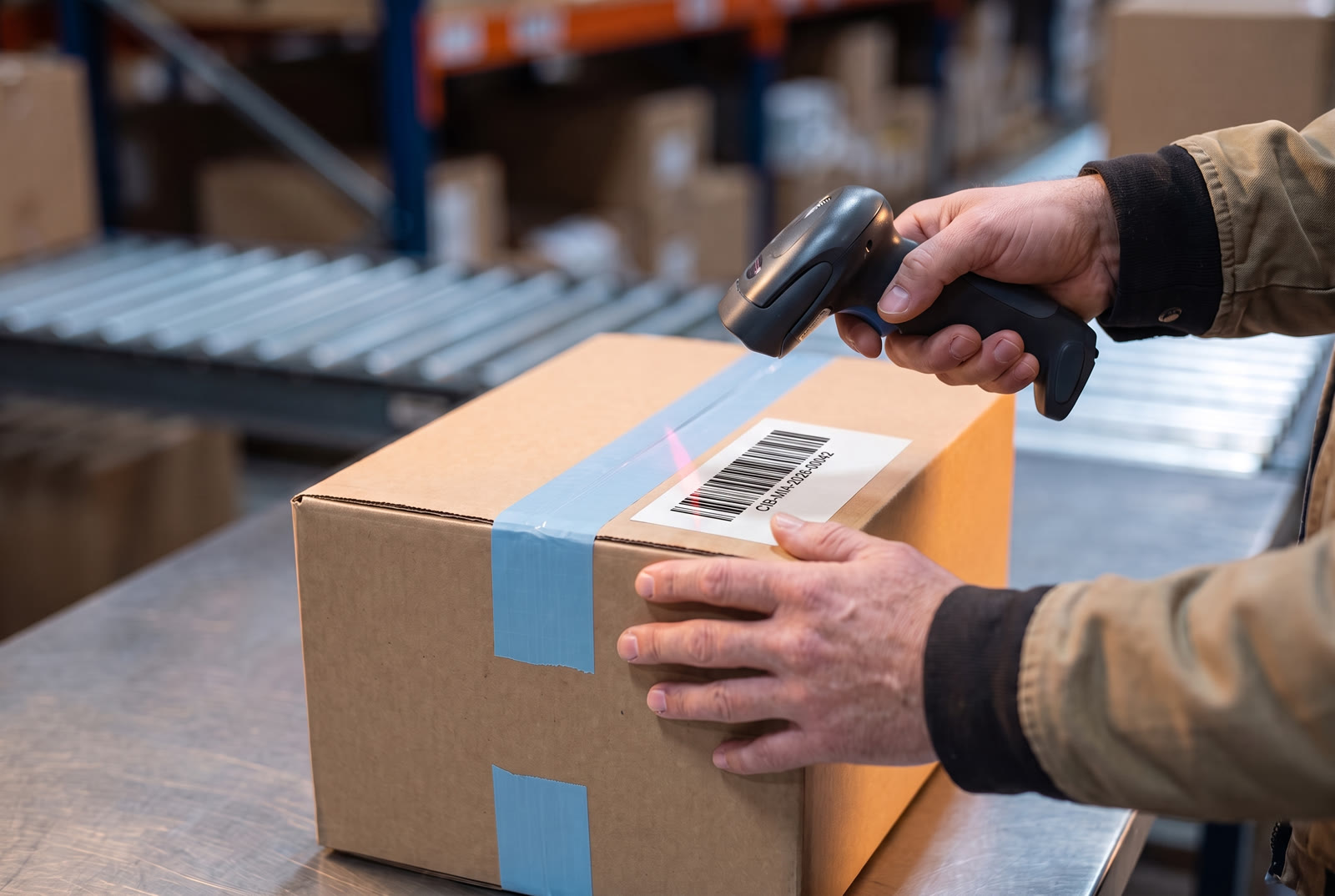 Hands scanning the shipping label barcode on a sealed kraft box with a handheld scanner at a warehouse dock